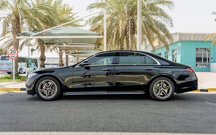 A black Mercedes S Class Qatar luxury sedan parked on a street near palm trees, with a turquoise building and a speed limit sign in the background on a sunny day.