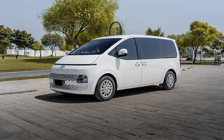 A white minivan, similar to the Mini Bus Vans Qatar style, is parked on a paved area in a park, surrounded by green trees and grass under a clear sky.