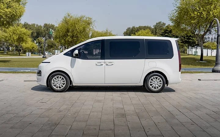 A white minivan, similar to the Mini Bus Vans Qatar fleet, is parked on a paved area in an open park, with green trees and grass in the background under a clear sky.
