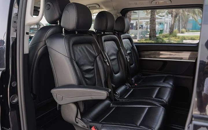 The image shows the interior of a Mercedes Viano in Qatar, featuring two rows of black leather seats with armrests, headrests, and seat belts. Large windows reveal palm trees and buildings outside.