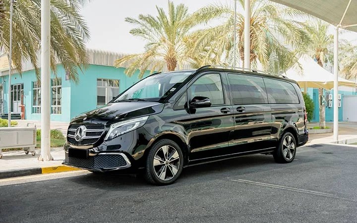 A black Mercedes Viano in Qatar is parked on a street under a canopy, with palm trees and modern teal buildings in the background.