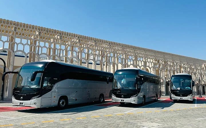 Three modern silver and black Premium Coach Bus Qatar vehicles are parked in a row on a paved area in front of a decorative arched building under a clear blue sky.