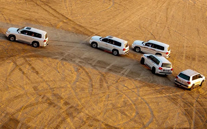 Five white SUVs drive in formation across sandy desert terrain during a デザートサファリ, leaving visible tire tracks and patterns in the golden sand as the サンライズ paints the horizon.