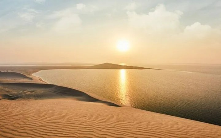 Golden sand dunes lead down to a calm body of water, reflecting the setting sun. An island rests in the distance beneath soft clouds, creating a peaceful scene—perfect for a 半日ツアー or デザートサファリ at sunrise.