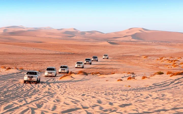 Seven white SUVs drive in a line across sandy desert dunes under a clear blue sky, their tire tracks and sparse vegetation visible—an exhilarating Desert Safari Excursion with Meal in Qatar.