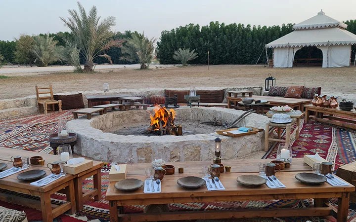 An outdoor seating area with rugs, wooden tables set for a meal, and cushions surrounds a large circular stone fire pit with a lit fire. Palm trees and a white tent in the background offer an authentic Qatari Farm Experience.