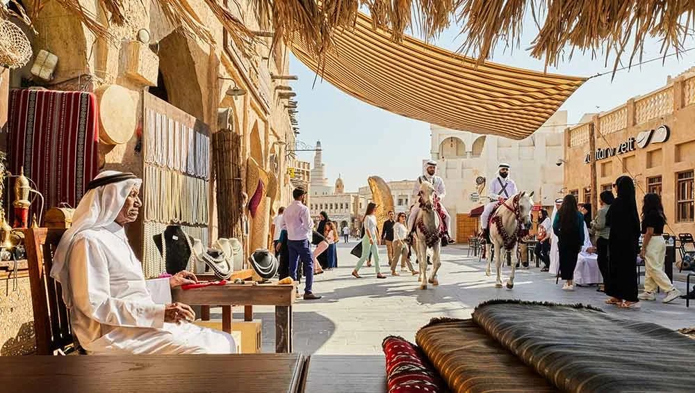A traditional Middle Eastern market scene captures Qatar’s heritage, with a man in white attire at a stall of woven goods, people strolling past, and two men riding horses through the sunlit street.