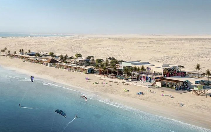 Aerial view of Fuwairit Beach Qatar, where a sandy shore lined with colorful buildings and palm trees meets the blue ocean. Several people are kite surfing near the coast, with a vast desert landscape stretching in the background.