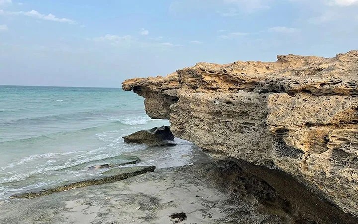 Large, jagged rock formation juts out over the sandy shores of Fuwairit Beach Qatar, framed by gentle blue-green waves and a partly cloudy sky.