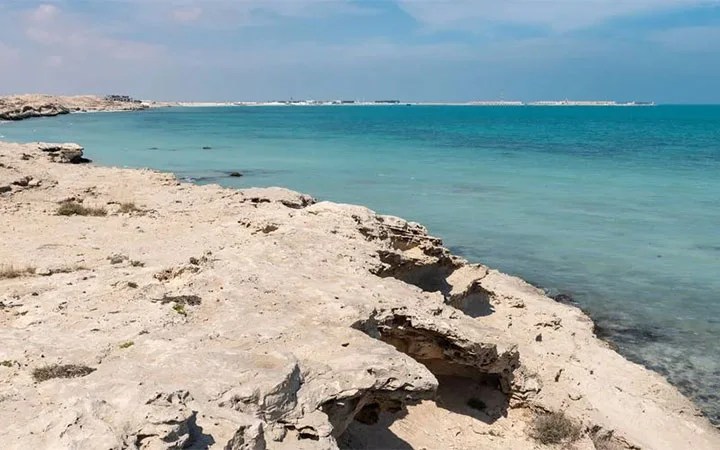 Rocky shoreline with light beige rocks meeting clear turquoise water under a partly cloudy sky at Fuwairit Beach Qatar, with a distant horizon visible across the calm sea.