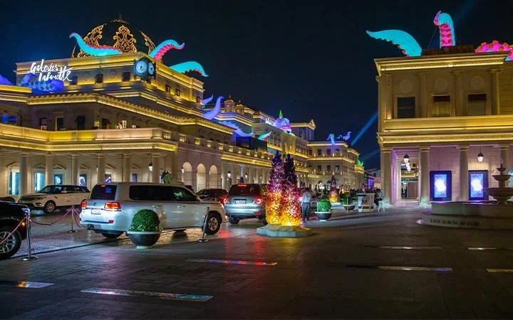 A night scene of the grand, illuminated Galeries Lafayette building with colorful tentacle sculptures on the rooftops, several parked cars, and decorative lights reflecting on the pavement.