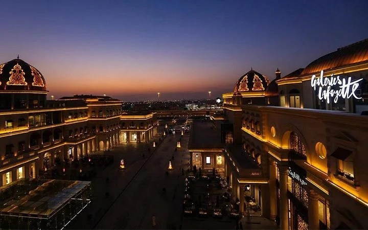 Open-air shopping mall at dusk, illuminated by warm lights. The iconic Galeries Lafayette sign glows atop a domed building to the right as the sky shifts from orange to deep blue with the coming night.