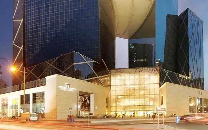 Modern glass and steel skyscraper with a unique curved design, illuminated at dusk near The Gate Mall Qatar. The lower level features large glass windows and a brightly lit entrance, with cars and streetlights visible in the foreground.
