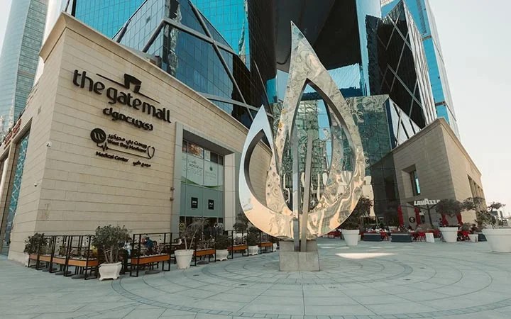 A large metallic flame-shaped sculpture stands in an open plaza outside The Gate Mall Qatar, surrounded by modern glass buildings and inviting outdoor seating areas on a sunny day.