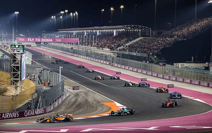 Formula 1 Qatar cars race on a brightly lit circuit at night, with grandstands full of spectators and a Fly to Doha sign overhead. The track features sharp turns, safety barriers, and prominent Qatar Airways branding.