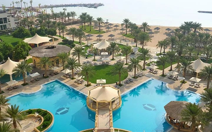 Aerial view of Intercontinental Doha, a luxury resort with a large blue swimming pool, palm trees, sun loungers, white canopies, and landscaped gardens, set along the sandy Doha Beach with calm sea in the background.