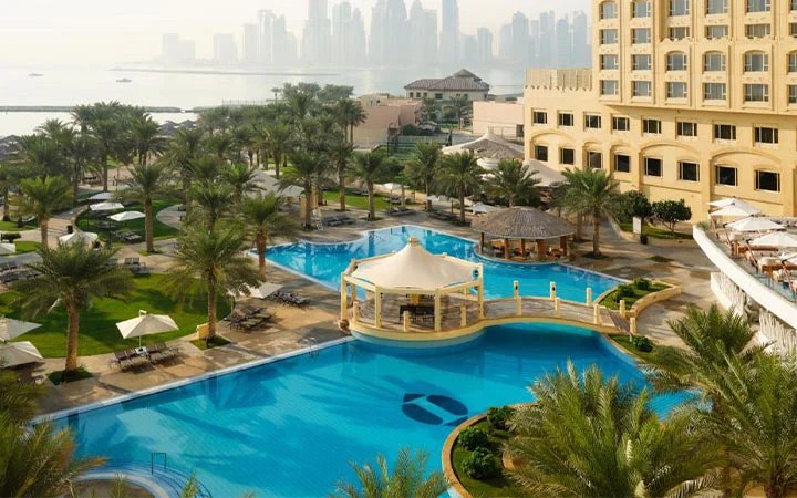 Luxurious hotel pool area at Intercontinental Doha Beach with palm trees, sun loungers, and shaded pavilions, overlooking a waterfront city skyline and tall buildings in the background on a hazy day.