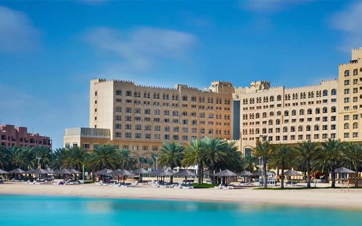 A large, luxurious hotel with a castle-like design stands behind rows of palm trees and beach umbrellas along the clear, turquoise shoreline of Intercontinental Doha Beach under a bright blue sky.