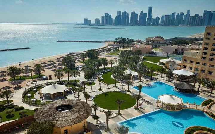 Aerial view of the Intercontinental Doha, a luxury beachfront resort with pools, palm trees, and cabanas on Doha Beach, overlooking a calm blue sea and the distant modern city skyline under a clear sky.