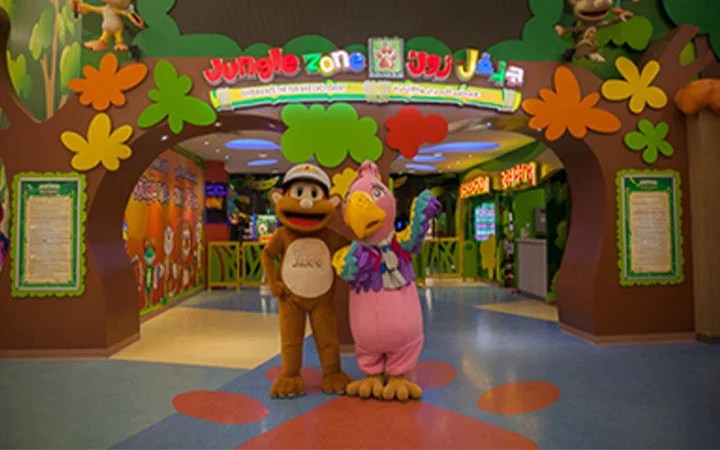 Two people in colorful animal costumes, a monkey and a bird, stand in front of the brightly decorated entrance to Jungle Zone, an indoor childrens amusement area with colorful signs and playful designs.