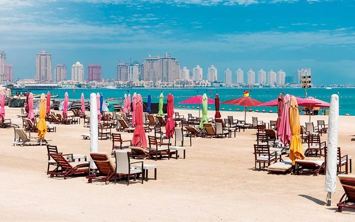 Colorful closed beach umbrellas and empty lounge chairs line the sandy shore of Katara Beach in Doha, with calm blue water and Qatar’s skyline of tall buildings in the background under a partly cloudy sky.