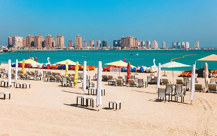 White sand beach with rows of chairs and umbrellas facing the turquoise sea at Katara Beach in Doha, Qatar; colorful paddle boats and kayaks are lined up near the water, with a city skyline under a clear blue sky.