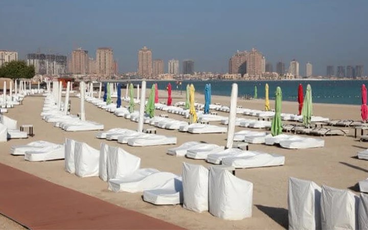 A sandy beach lined with empty white lounge chairs and closed umbrellas in various colors at Katara Beach, facing calm blue water, with Doha’s striking city skyline of tall buildings in the background.