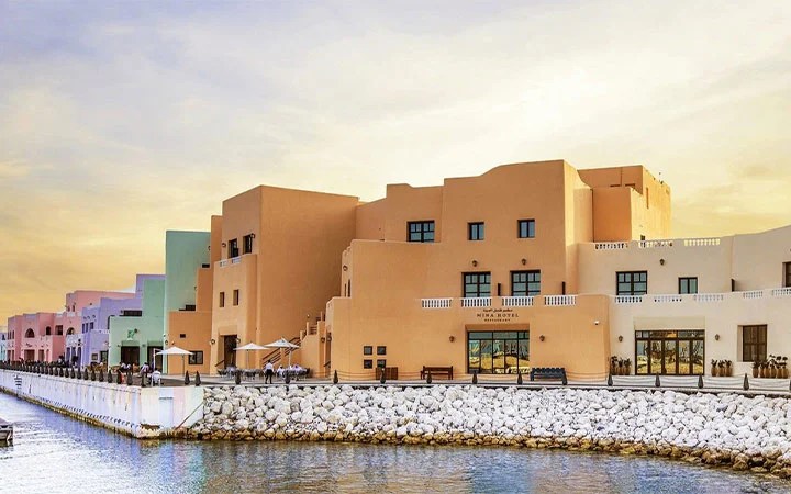 Colorful, modern buildings line a waterfront promenade with rocky barriers and calm water in the foreground, set against a soft, golden sky at sunset.