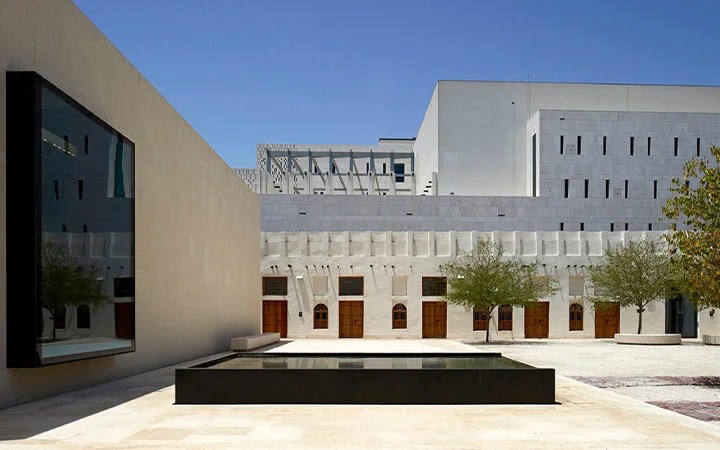 A modern courtyard at Msheireb Museums in Doha, Qatar, features minimalist architecture with light stone walls, a rectangular reflecting pool, neatly planted trees, and wooden doors under a clear blue sky.