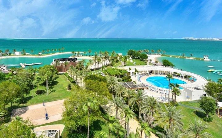 A lush beachfront resort, Sheraton Doha Beach, features a circular pool, palm trees, and green lawns beside a turquoise sea under a bright blue sky, creating a tropical vacation atmosphere.