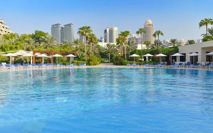 A large outdoor swimming pool at Sheraton Doha Beach is surrounded by lounge chairs, umbrellas, palm trees, and lush greenery, with city skyscrapers visible in the background under a clear blue sky.