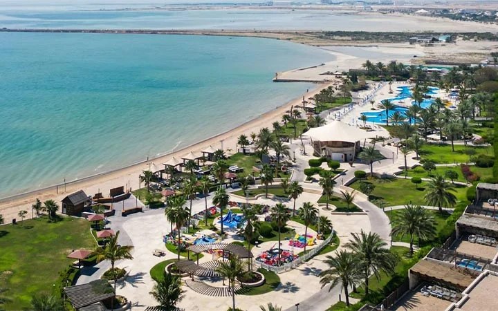 Aerial view of Simaisma Beach resort in Qatar, with palm trees, swimming pools, green lawns, lounge chairs, shaded huts, and a sandy shoreline beside the calm blue sea under a clear sky.