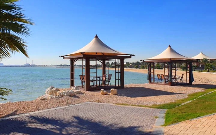 Two wooden gazebos with tables and chairs stand on the sandy shore of Simaisma Beach in Qatar, beside calm blue water under a clear sky, with palm trees and green grass nearby.