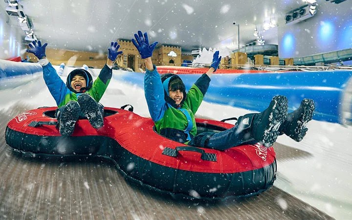 Two people in winter clothing smile and raise their arms while sliding down a snowy indoor slope together in connected red snow tubes, with artificial snowflakes falling around them.