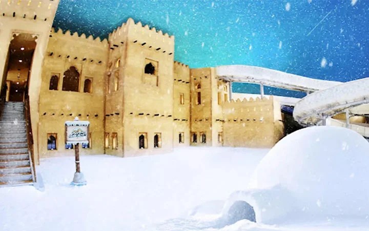 A large sand-colored fortress building stands under a bright blue sky, surrounded by snow. Snow falls gently, covering the ground and an igloo in the foreground. A sign and stairway are visible on the left.