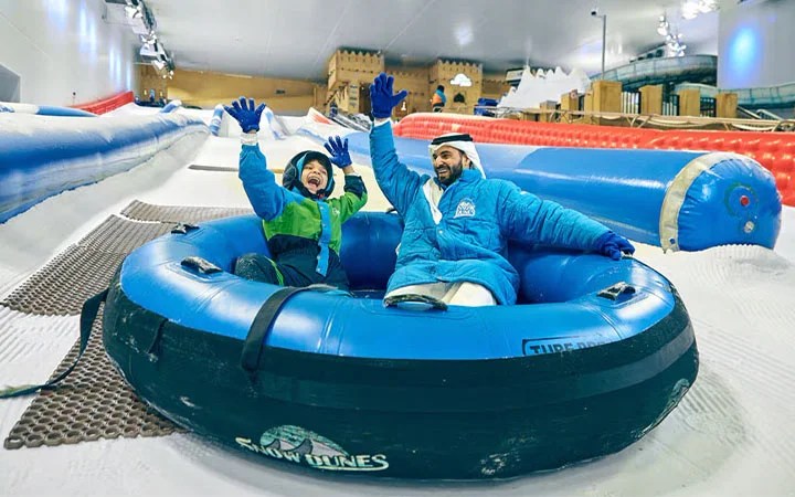 Two people in winter clothes smiling and waving as they sit together in a large blue snow tube on an indoor snow slide, surrounded by artificial snow and other tubing lanes.