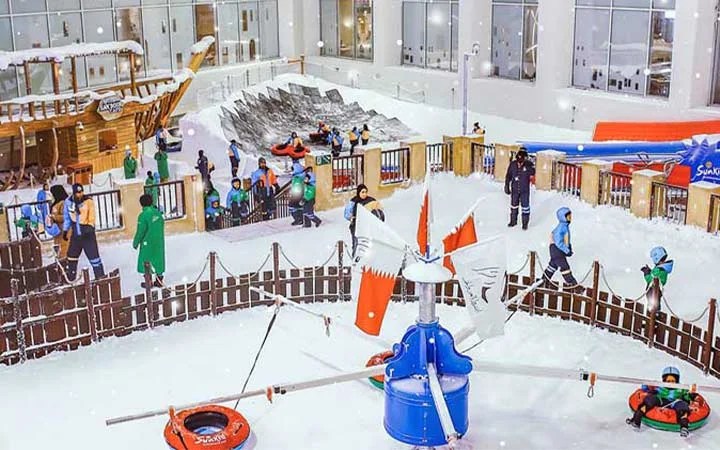 Children and adults enjoy various winter activities in an indoor snow park, featuring snow slides, a wooden ship, snow-covered ground, and Bahraini flags, with people dressed in winter clothing.