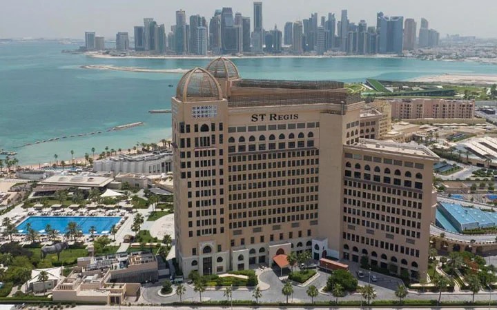 A large luxury hotel, St. Regis, stands near the waterfront with lush gardens, a pool, and exclusive access to St Regis Beach. In the background, modern skyscrapers rise across the bay under a clear sky.