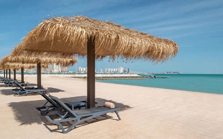 Rows of lounge chairs with straw-thatched umbrellas line the sandy shores of St Regis Beach, beside calm turquoise water, with a city skyline visible in the background under a clear blue sky.