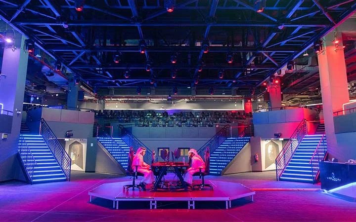 Two people sit facing each other at a gaming table in the center of a brightly lit, futuristic arena with blue and pink lights illuminating staircases and seating in the background.