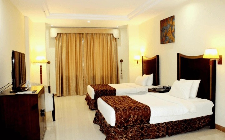 Enjoy a comfortable 3-star stay in this Tourist Hotel room featuring two single beds with white linens and brown runners, a TV on a wooden desk, wall lamps, large beige curtains, and a painting above the beds.