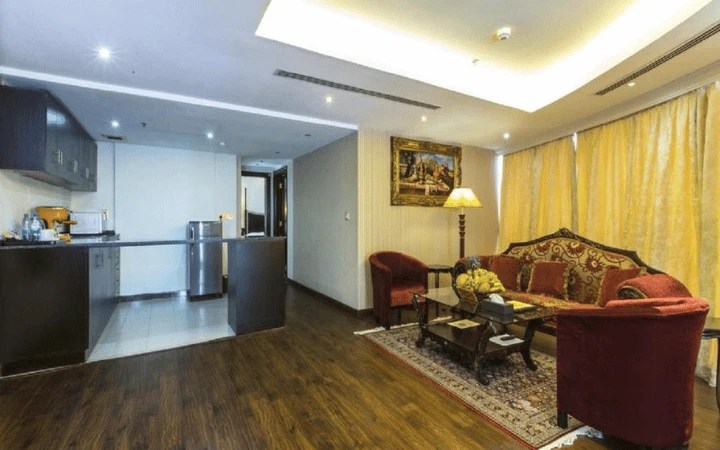 Modern Tourist Hotel suite with a small kitchen featuring dark cabinets and a stainless steel fridge; cozy living area with red armchairs, a patterned sofa, glass coffee table, and gold curtains—perfect for a 3-star stay.