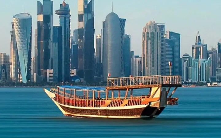 A traditional wooden boat floats on calm blue water with a modern city skyline of tall, glass skyscrapers in the background—perfect for an unforgettable Iftar Cruise experience.