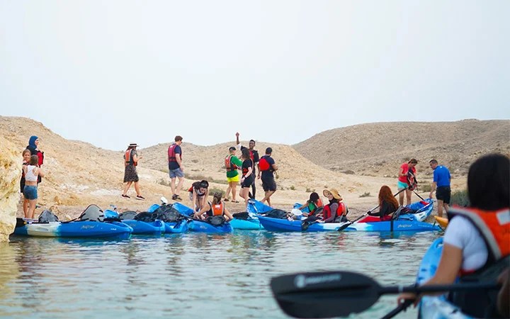 A group of people wearing life jackets gather near blue kayaks by a rocky shoreline, preparing for kayaking. Some enjoy the calm water while others share an iftar meal together, with arid hills rising in the background.