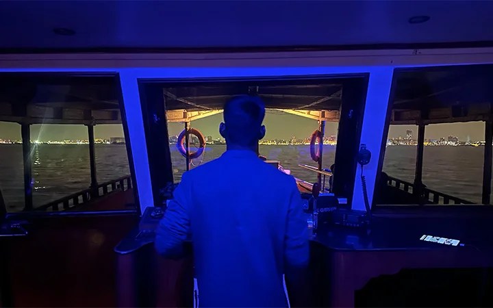 A person stands at the helm of a private dhow boat at night, gazing over dark water toward a brightly lit city skyline in the distance, illuminated by blue interior lights—a serene scene perfect for a Ramadan offer.