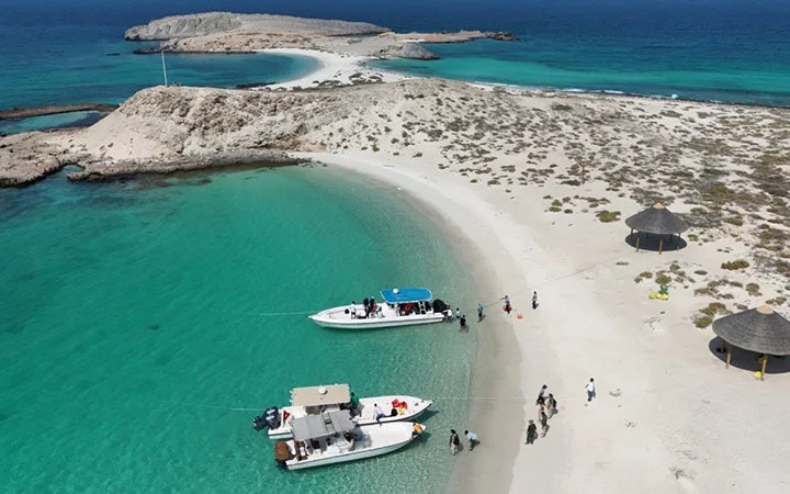 Aerial view of Awa Island’s sandy beach with turquoise water, three luxury boats anchored near shore, people strolling along the beach, and a small island connected by a sandbar—the perfect Day-Cation escape.