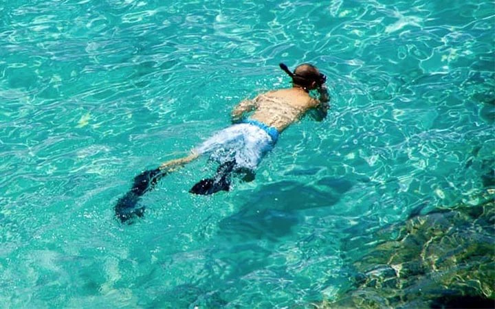 A person wearing snorkeling gear, including a mask and fins, floats face-down in clear turquoise water near the rocky edge of Shura Awa Island, enjoying a tranquil Luxury Day-Cation.