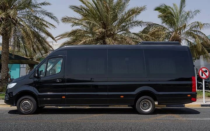 A large black Mercedes Sprinter VIP Bus is parked on a street lined with palm trees in Doha. There is a no-right-turn traffic sign visible in the background. The scene appears sunny and clear.