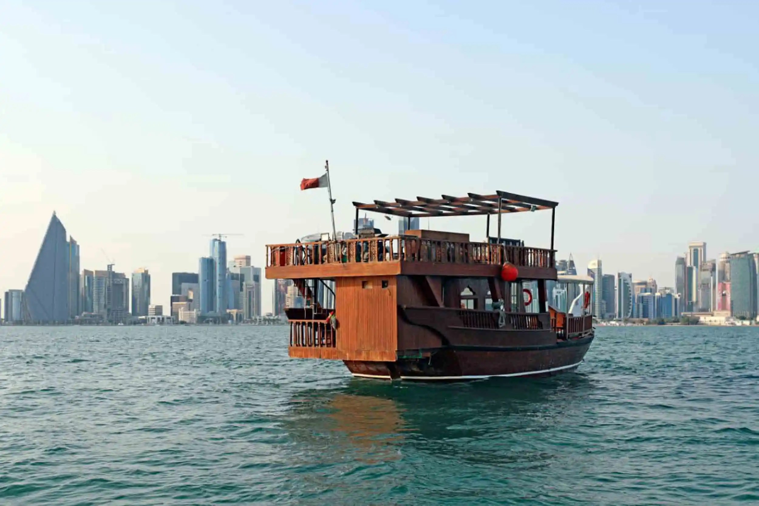 A traditional wooden dhow boat floats on the water with a city skyline and modern skyscrapers in the background under a clear sky.