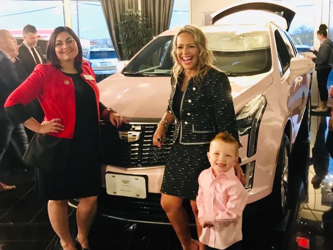 “Group of people posing in front of a light pink Cadillac SUV inside a showroom—celebrating achievement, legacy, and Mary Kay success.”
